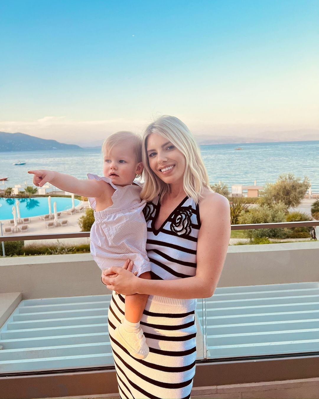 a woman in a striped dress is holding a baby and pointing at the ocean