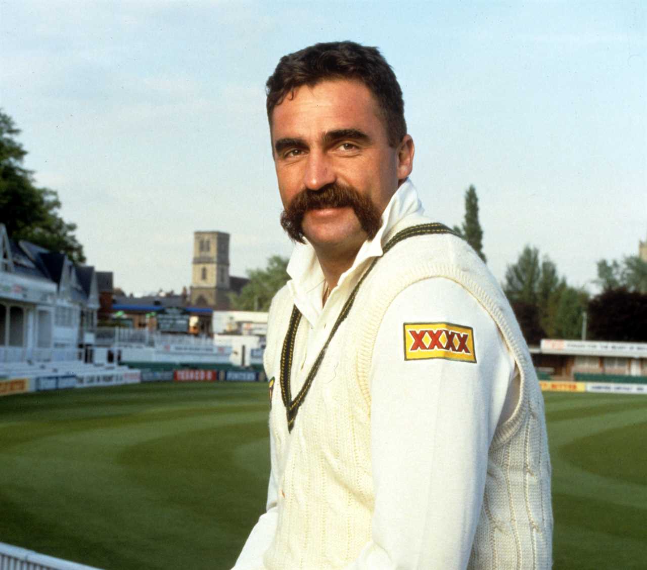 a man with a mustache wears a white vest with a xxx logo on it