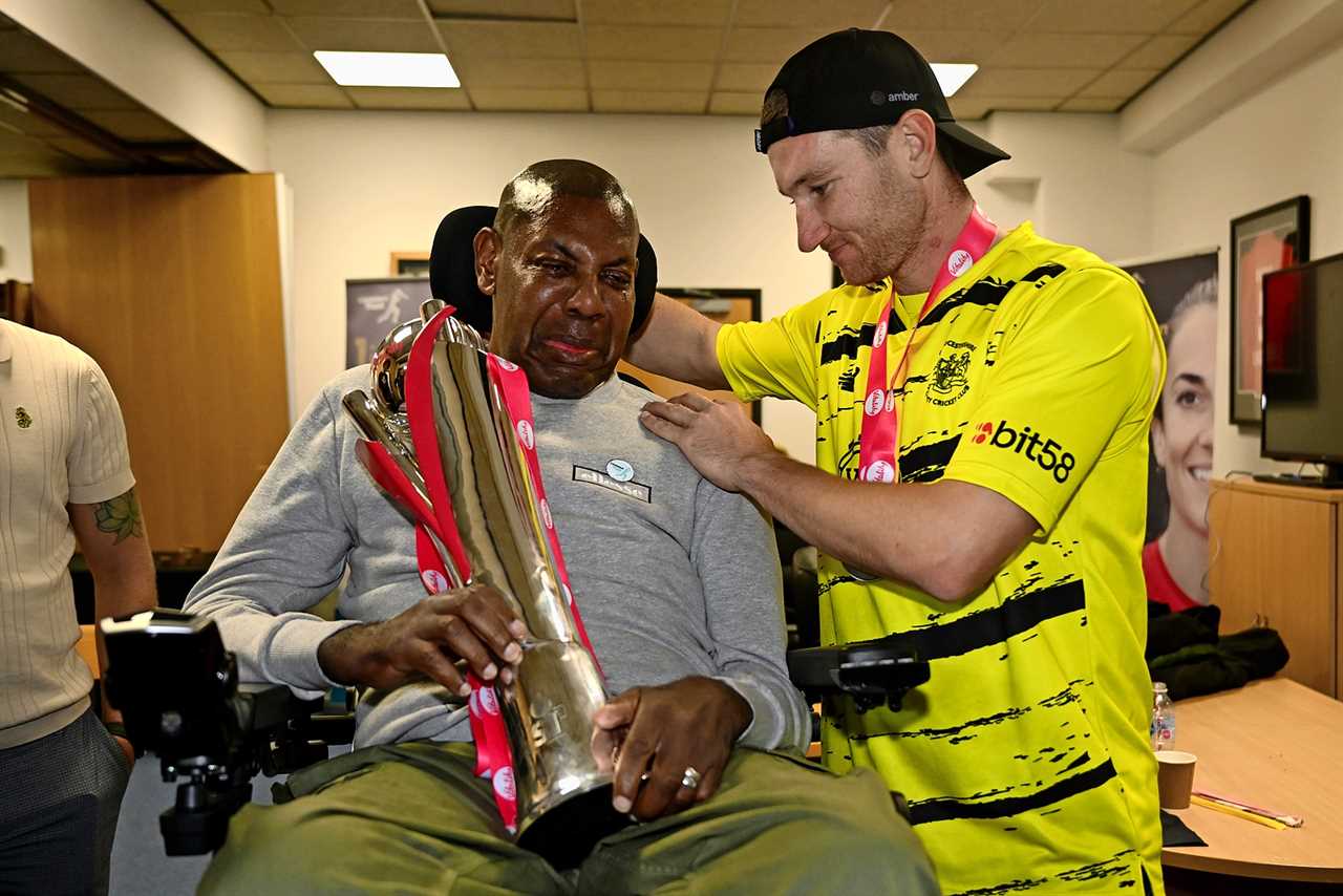 A cricket player presents a trophy to another man in a wheelchair.