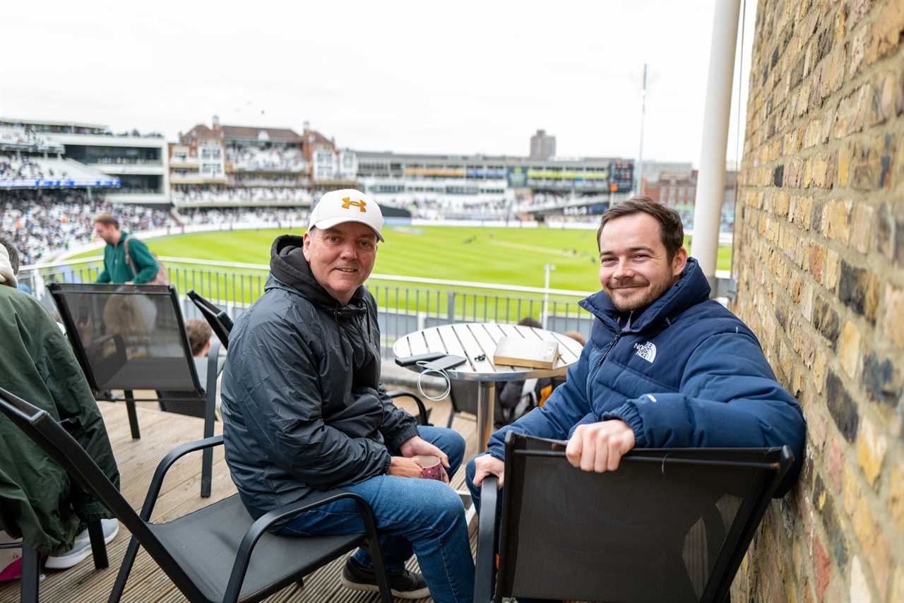 Two men sitting on a balcony overlooking a cricket field.